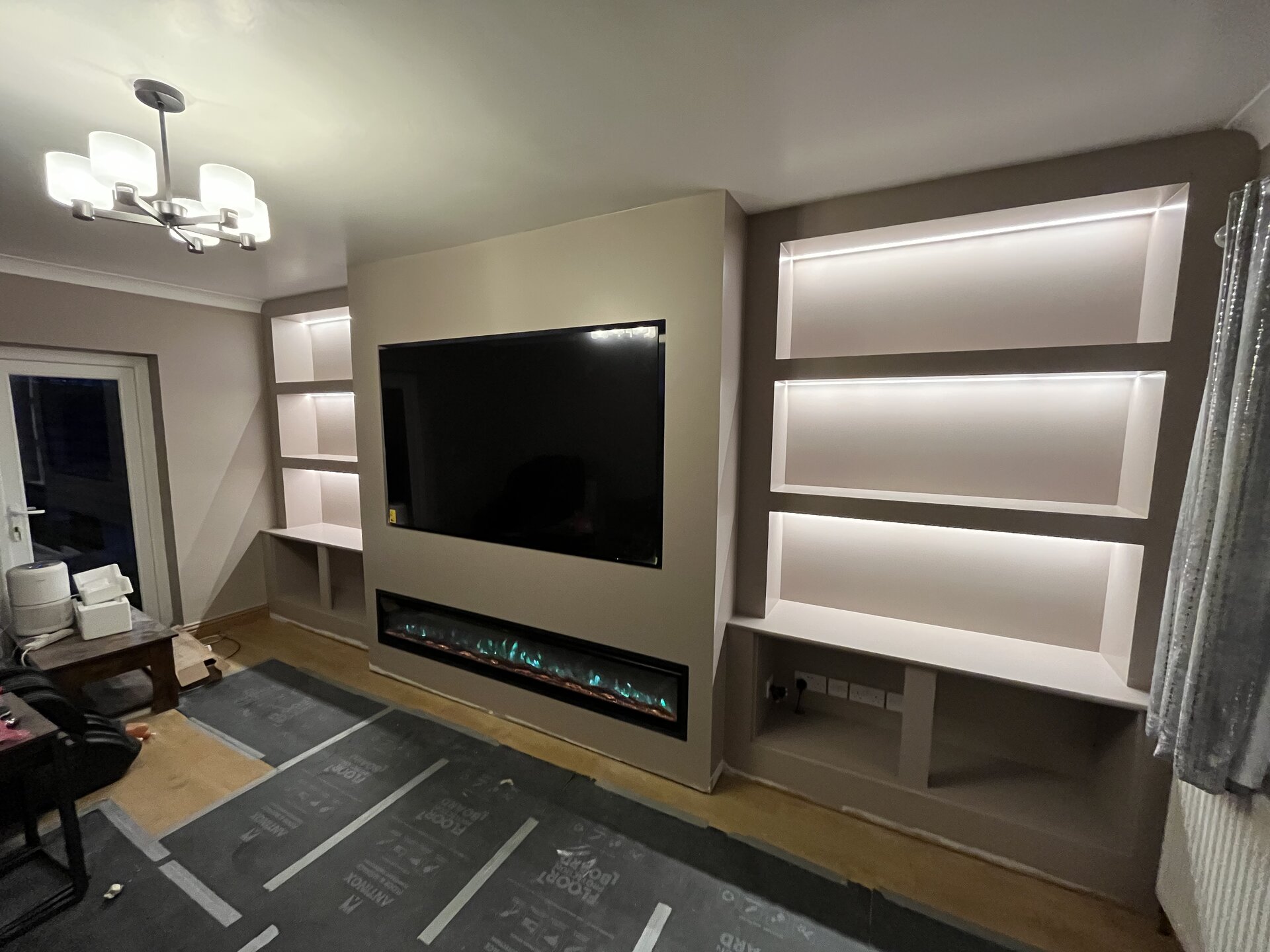 Near-finished bespoke media wall with electric fireplace lit and warm LED alcove shelving in Sawtry