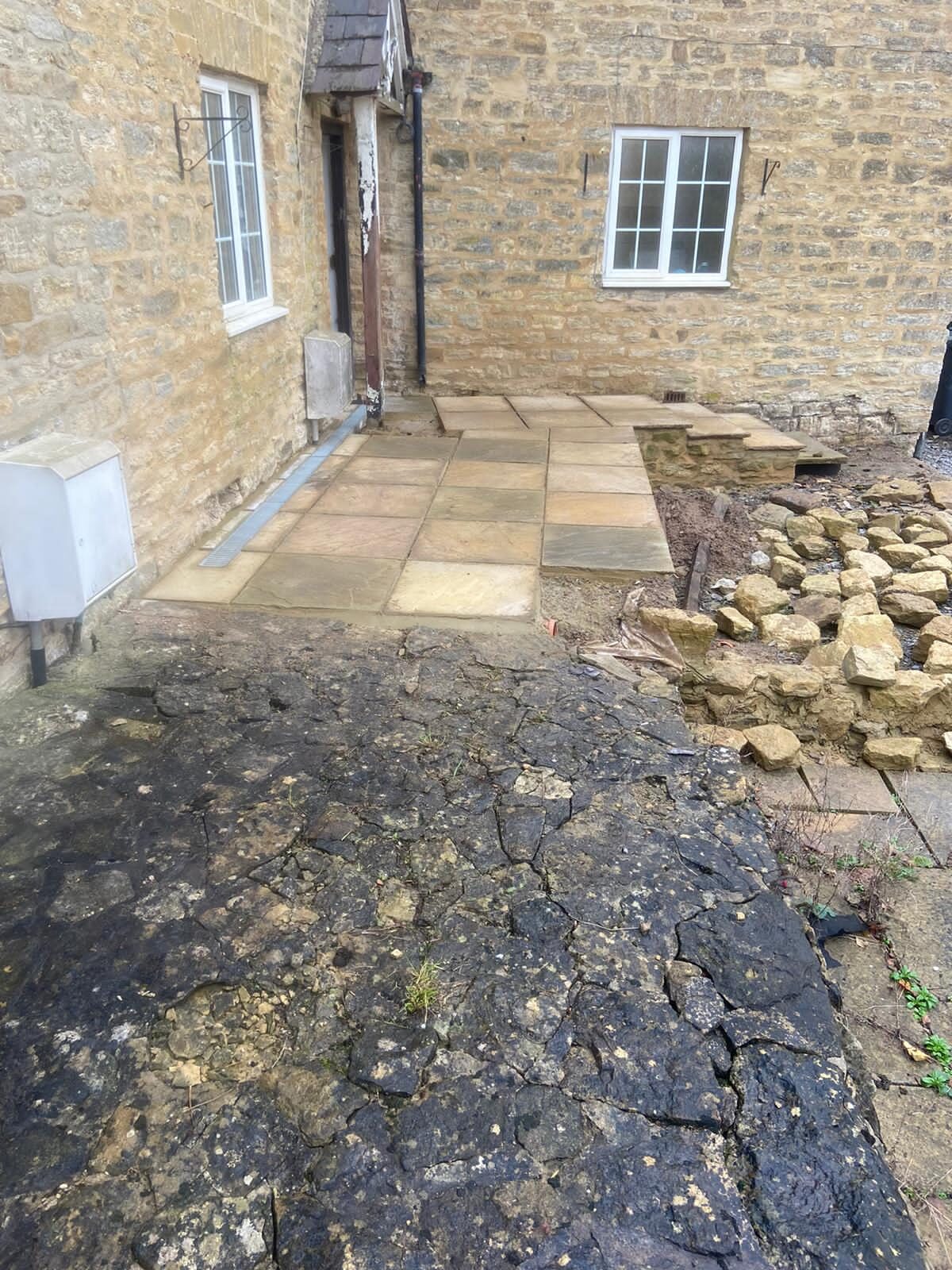 Reclaimed sandstone path being laid against rear cottage door beside broken tarmac in Turvey, Bedfordshire